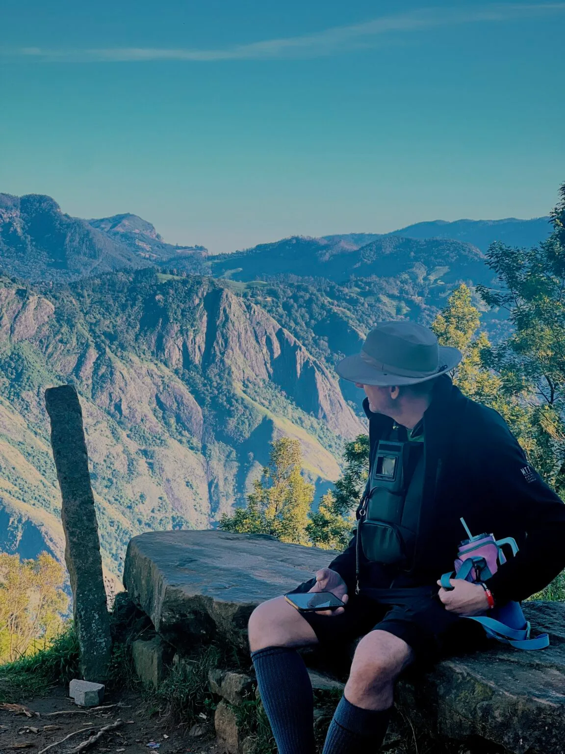 A guest sits on a rock at the edge of a cliff, looking out over forested mountains