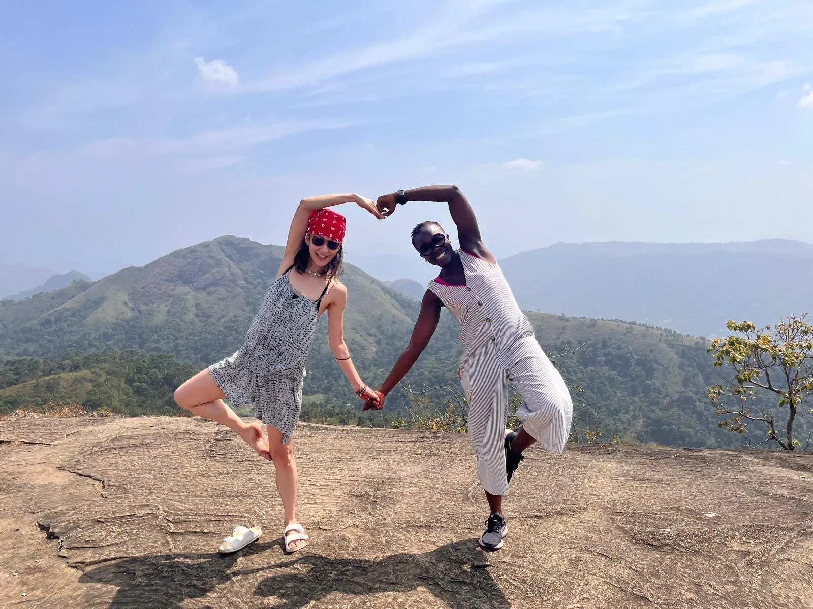 Two guests balance in tree pose and make a heart with their arms on a hilltop