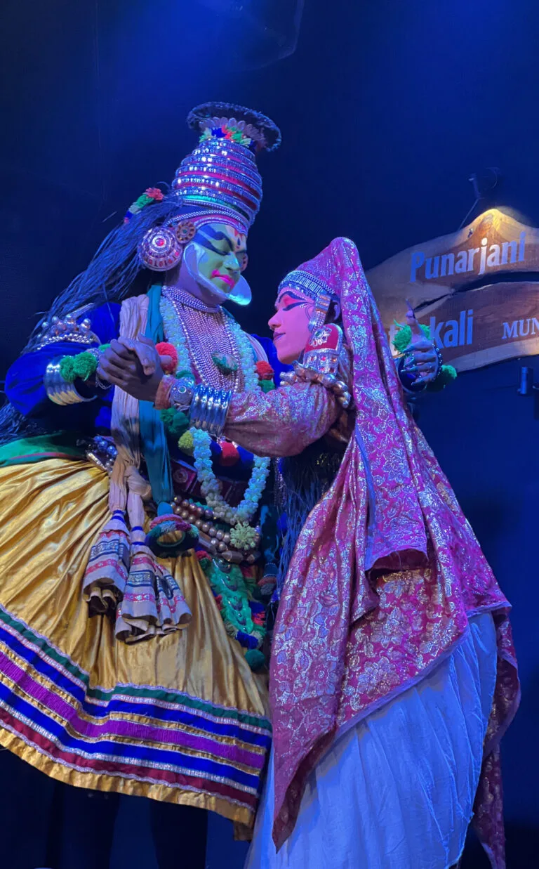 Two Kathakali performers in painted faces and ornate costumes on stage
